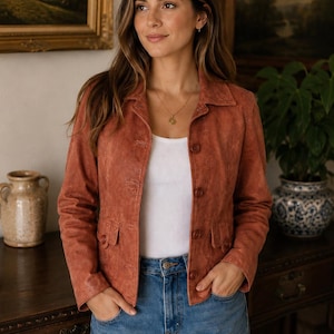 May include: A rust-coloured suede jacket with a button-up front and two front pockets. The jacket is paired with a white top and blue jeans. The jacket has a classic, tailored design.