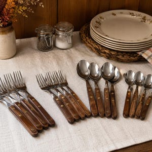 May include: A flatware set consisting of stainless steel forks and spoons with dark brown wooden handles secured by two metal rivets. The collection includes multiple forks and spoons arranged on a light-colored fabric surface, featuring a classic design.