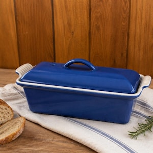 Puede incluir: Un molde rectangular de cerámica para pan con un acabado azul profundo brillante y una tapa a juego. El molde cuenta con asas laterales integradas con textura acanalada y un borde interior blanco, presentado junto a pan rebanado y una ramita de romero fresco.