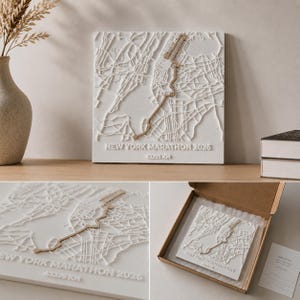 Könnte beinhalten: Eine weiße quadratische Wandplatte mit einer erhabenen 3D-Karte der New York Marathon-Strecke in Beige. Der Text NEW YORK MARATHON 2026 und 42,195 KM ist unten eingeprägt. Das Stück ist ausgestellt und in einem Versandkarton zu sehen.