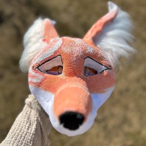 May include: A plush fox mask with orange, white, and black details. The mask has large, expressive eyes, a black nose, and white fur around the ears. The fox's face is held by a person wearing a beige sweater.