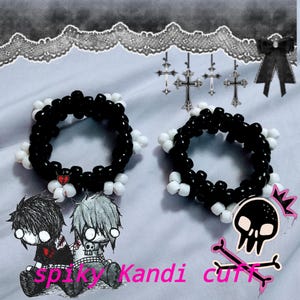 Può includere: Due braccialetti kandi di perline bianche e nere con un ciondolo a forma di cuore. L'immagine include una grafica di teschio e ossa incrociate, personaggi dei cartoni animati e il testo "spiky Kandi cuff."