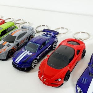 May include: A selection of toy car keyrings in various colours, including red, blue, and grey. The keyrings feature detailed car designs with racing stripes and logos. Each car is attached to a silver metal keyring.