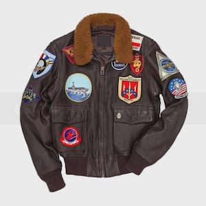 May include: A brown leather bomber jacket with a brown faux fur collar. The jacket features multiple embroidered patches, including a ship, a Tomcat patch, and the text "SEABEES". It has two front pockets and a ribbed hem and cuffs.