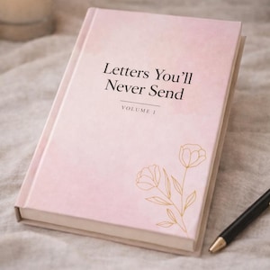 Puede incluir: Un diario rosa titulado "Letters You'll Never Send, Volume 1" con un diseño floral dorado. Un bolígrafo negro descansa sobre una superficie suave de color neutro, con una vela encendida en el fondo. La portada del diario tiene un efecto acuarela.