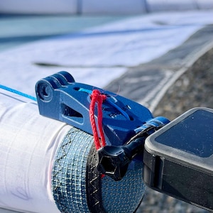 May include: Close-up of a blue and white kite surfing sail with a blue plastic component and a red rope. A black device is attached to the sail. The sail is on a gravel surface.