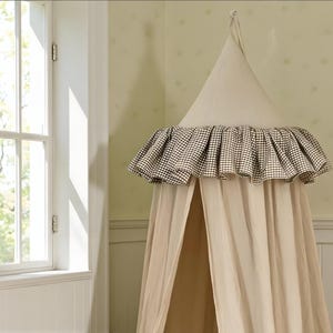 May include: A beige canopy bed with a cone-shaped top and a ruffled trim in a black and white checkered pattern. The canopy hangs from the ceiling, creating a cozy and enclosed space. The bed is in a room with a window.