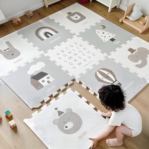 Könnte beinhalten: Grau-weiße, ineinandergreifende Spielmatte mit Illustrationen von Tieren, einem Regenbogen, einem Haus, einem Heißluftballon, einem Baum und dem Alphabet. Ein Kind spielt auf der Matte.