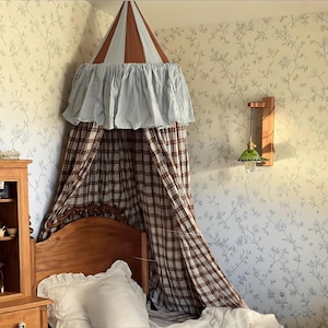Puede incluir: Una cama con dosel con una cortina a cuadros marrón y blanca y una parte superior a rayas azul claro y marrón. La cama tiene una cabecera de madera y una almohada blanca. La habitación tiene papel pintado floral y una lámpara de cristal verde.