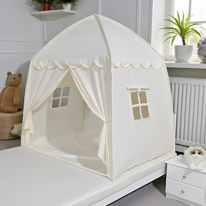 May include: A cream-colored playhouse tent with scalloped trim and two window cutouts. The tent is set up on a white mattress, creating a cozy indoor play space for children. The tent has tie-back curtains.