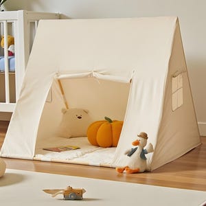 May include: A cream-coloured play tent with an A-frame design. Inside, a white bear-shaped cushion and an orange pumpkin-shaped cushion are visible. A stuffed duck toy with a hat and vest is in front of the tent. A small wooden toy aeroplane is on the floor.
