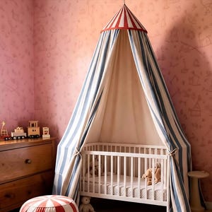 May include: A white wooden crib positioned under a hanging fabric canopy featuring vertical blue and white stripes with a red and white striped conical top. The canopy drapes down to the floor on both sides, creating a tent-like structure.