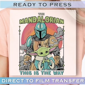 May include: A light pink t-shirt featuring a graphic of The Mandalorian and Grogu, with the text "THE MANDALORIAN" and "THIS IS THE WAY". The design includes a helmeted figure and a green alien against a sunset backdrop.