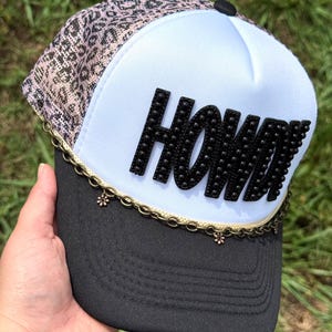 Puede incluir: Gorra de camionero negra y blanca con la palabra "HOWDY" en letras negras con tachuelas. La parte superior de la gorra tiene un estampado de animales rosa y negro, y la visera es negra. Una cadena dorada con dijes de flores decora la visera.