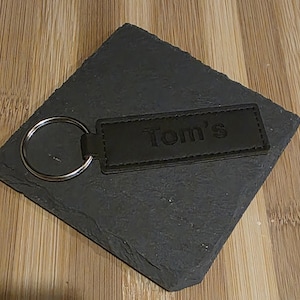 Puede incluir: Un llavero de cuero negro con un llavero plateado. El llavero es rectangular y tiene el nombre "Tom's" grabado en relieve. El llavero descansa sobre un posavasos cuadrado gris oscuro, colocado sobre una superficie de madera.