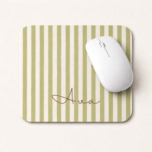 May include: A white wireless computer mouse rests on a square mousepad. The mousepad has a green and white striped pattern with the name "Ava" in a brown script font. The mousepad is designed for computer use.