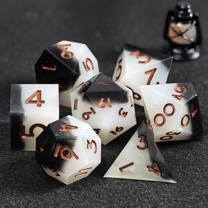 May include: A set of seven polyhedral dice featuring a blended black and white resin design with metallic copper-colored numbers. The collection includes various geometric shapes such as a d20, d12, d10, d8, d6, and d4, all displaying clear numerals.