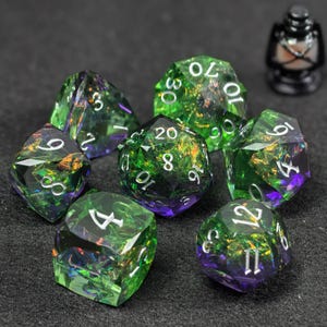 Puede incluir: Un juego de siete dados poliédricos con un degradado translúcido verde y morado con escamas de papel iridiscente incrustadas. Cada dado tiene bordes afilados y números grabados en blanco, incluyendo d20, d12, d10, d8, d6, d4 y un dado porcentual.