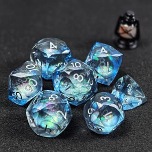 Può includere: Un set di sette dadi poliedrici realizzati in resina blu traslucida con scaglie di glitter iridescenti all'interno e numeri color argento. I dadi presentano bordi affilati e sfaccettati e sono disposti in gruppo con una piccola lanterna nera.