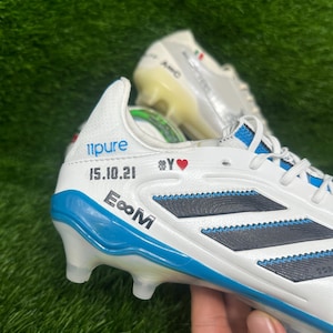 Può includere: Scarpini da calcio bianchi e blu con strisce nere e il testo "11pure", "15.10.21" e "E∞M". Gli scarpini sono su una superficie verde.