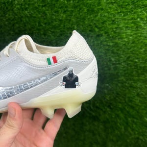 May include: White and silver soccer cleat with a textured design and a clear sole. Features a small sticker of a person in a black suit and an Italian flag. The cleat is held against a green background.