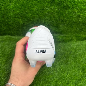 Può includere: Scarpino da calcio bianco e rosa con la scritta "ALPHA" stampata sul tallone. Lo scarpino ha tacchetti trasparenti e la scritta "AVENGER" è stampata sul lato. Lo scarpino è tenuto su uno sfondo di erba sintetica verde.
