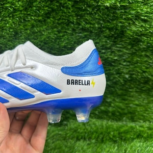 Puede incluir: Bota de fútbol blanca y azul con el nombre "BARELLA" y un rayo. La bota tiene una franja azul y un contrafuerte azul con el logo de Adidas. La suela tiene tacos transparentes.