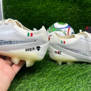 Puede incluir: Un par de botas de fútbol blancas con detalles plateados y un diseño texturizado. Las botas tienen una suela clara y presentan la palabra "SHQIRI" y un gráfico de alienígena. Un balón de fútbol es visible en el fondo.