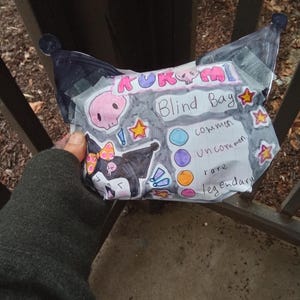 May include: A handmade Kuromi blind bag, featuring a black and white cartoon character with a pink bow. The bag includes a pink skull and star stickers, and the text "Blind Bag" with rarity indicators: common, uncommon, rare, and legendary.