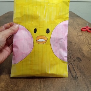 Puede incluir: Una bolsa de papel amarilla hecha a mano con una cara de pato de dibujos animados dibujada. El pato tiene ojos negros, un pico naranja y mejillas rosadas. La bolsa está sostenida por una mano y descansa sobre una superficie de madera.