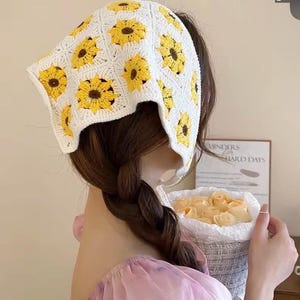 May include: A white crochet headscarf featuring yellow sunflower designs. The head covering is square-shaped and worn on the head. The sunflowers have yellow petals and brown centers. The headscarf is handmade and has a vintage aesthetic.