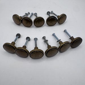 Könnte beinhalten: Ein Set aus elf runden Schrankknöpfen mit dunkelbronzenem Finish. Jeder Knopf hat eine glatte, kreisförmige Vorderseite und eine Metallschraube, die an der Rückseite herausragt. Die Knöpfe sind in zwei Reihen vor einem hellen Hintergrund angeordnet.