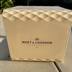 Puede incluir: Cubo de hielo para champán de color crema con un patrón de diamantes en el borde superior. La parte frontal del cubo presenta el logotipo "MOËT & CHANDON CHAMPAGNE" en negro, con una corona y una estrella. El cubo está sobre una superficie de hormigón.