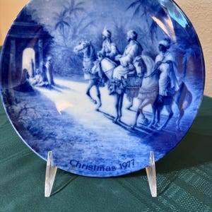 May include: A round ceramic decorative plate in a deep blue and white color palette. The illustration depicts three figures riding horses toward a structure with a glowing doorway. The text "Christmas 1977" is printed in blue at the bottom edge.