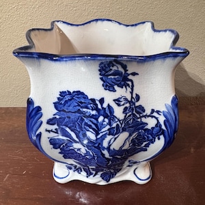 May include: A square ceramic planter with a scalloped rim and a white base featuring a blue floral pattern. The surface has a visible crackle glaze finish. The planter stands on four small feet and displays deep blue rose motifs on the front.