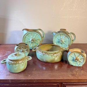 May include: A collection of six ceramic kitchen items featuring a light green and brown glaze. The set includes a pitcher, a teapot with a lid, a lidded sugar bowl, a large serving bowl with two handles, and two small open containers, all with wagon wheel motifs.