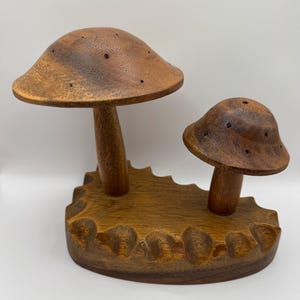May include: A wooden mushroom sculpture featuring two mushrooms of different sizes. The mushrooms are brown with small holes and sit on a carved wooden base. The sculpture is made of a warm-toned wood.