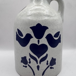 May include: A white ceramic jug with a speckled finish and a single handle. The front features a dark blue graphic design consisting of a central heart, a tulip above it, two birds facing inward, and two smaller floral motifs at the bottom.