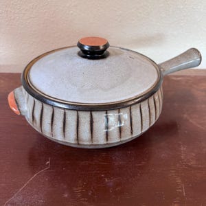 Puede incluir: Una cazuela de cerámica con tapa, que presenta un acabado gris claro moteado y ranuras verticales de color marrón oscuro en los lados. La tapa tiene un pequeño pomo naranja, y el plato incluye un asa lateral y un pequeño acento naranja.