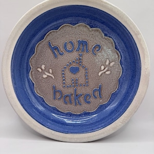 Può includere: Una ciotola rotonda in ceramica con bordo bianco e interno blu. Il centro presenta un cerchio beige testurizzato e smerlato con il testo blu "home baked" e il contorno punteggiato di una casa contenente un cuore. Due piccole foglie bianche.