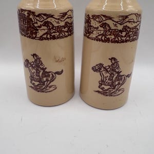 May include: A pair of cylindrical ceramic salt and pepper shakers in a light beige color. Each shaker features a dark brown printed design showing a rider on horseback and a stagecoach scene with horses traveling across a landscape.