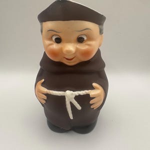 May include: A ceramic figurine of a monk, featuring a brown robe, a dark brown cap, and a white rope belt. The monk has rosy cheeks and a smiling expression. The figurine is standing upright.