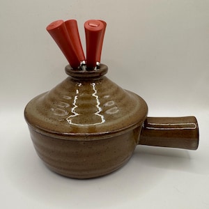 May include: A brown ceramic fondue pot with a lid and handle. The pot has a speckled finish and is filled with three red-handled fondue forks. The pot is designed for serving melted cheese or chocolate.