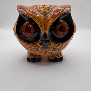 May include: A ceramic owl figurine featuring a glossy brown and tan glaze with a textured feather pattern. The owl has large, prominent dark brown eye sockets surrounding bright orange irises, a small black beak, and two small feet.