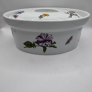May include: A white ceramic casserole dish with a lid, featuring a botanical pattern of purple flowers, green leaves, and small butterflies. The round vessel has a wide rim and a smooth, glossy finish, showcasing detailed floral illustrations.