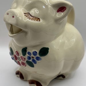 Op de afbeelding: Een crèmekleurige keramische kan in de vorm van een varken met een lachend gezicht en bloemaccenten. De kan heeft bruine wimpers, roze oren en bruine poten. De bloemen zijn roze, blauw en groen.