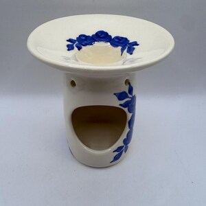 May include: A cream-colored ceramic oil burner featuring a wide, shallow top dish and a cylindrical base with a large arched opening. The piece is decorated with hand-painted blue floral motifs, including roses and leaves, on the top and side.