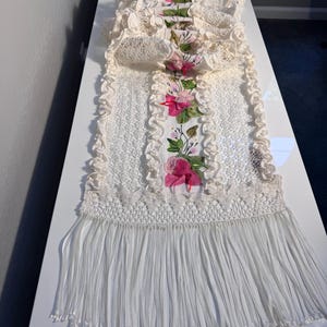 May include: A long, rectangular table runner featuring cream-colored crocheted lace borders with ruffled edges. The center panel is white fabric decorated with embroidered pink and red roses, green leaves, and long, flowing white fringe at both ends.