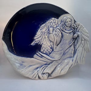 May include: A round ceramic decorative plate featuring a deep cobalt blue glazed center. A raised, three-dimensional white relief sculpture of an angel with flowing robes and large wings extends from the right side, partially overlapping the blue basin.