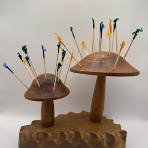 May include: Two wooden mushroom-shaped toothpick holders with a carved base. The holders are brown and feature colourful toothpicks. The toothpicks have green, blue, and yellow decorative tops. A whimsical addition to any kitchen or dining area.